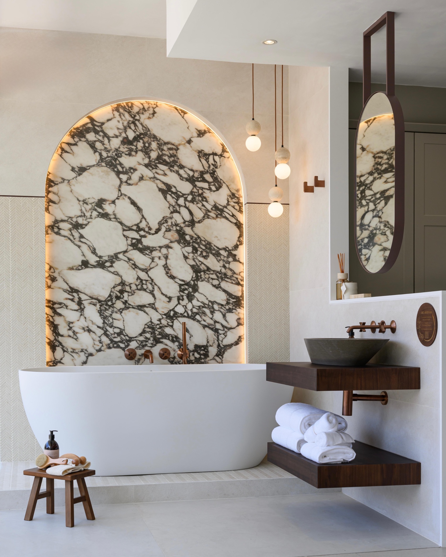 Bathroom by Clo Interior Designs with a freestanding bath beneath a backlit marble arch, walnut vanity and oval mirror